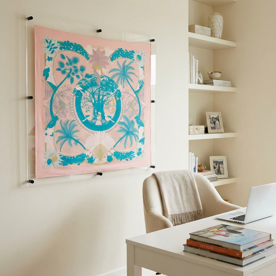 A pink silk scarf with a turquoise and white botanical and monogram pattern is displayed on a clear acrylic stand mounted on a beige wall in a modern home office setting.