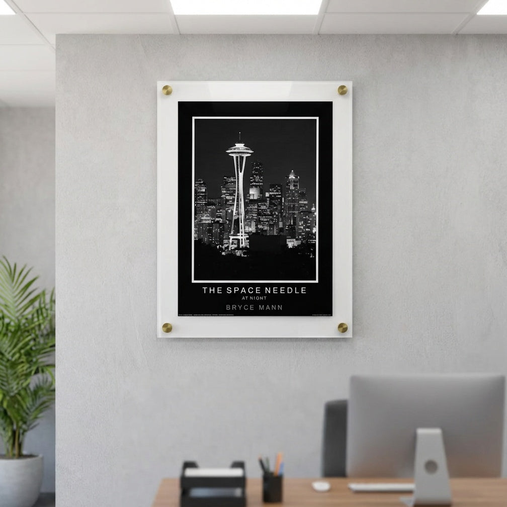A framed black and white poster of Seattle's Space Needle and skyline, mounted on a light gray wall above a wooden desk with a computer monitor and office accessories.