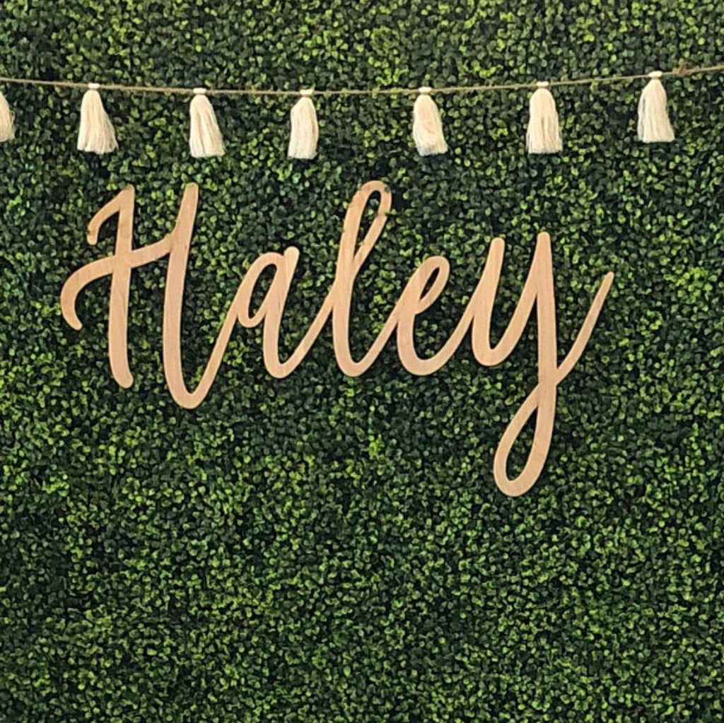 Custom wood name sign reading Haley in cursive script mounted on a green boxwood backdrop with tassel garland