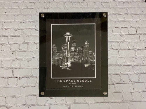 Black floating acrylic picture frame holding a Space Needle photo print on a white brick wall